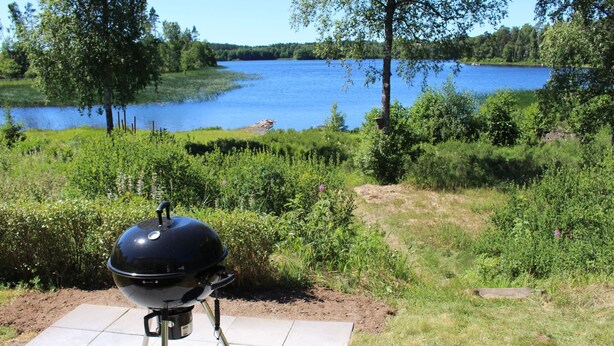 Una parrilla de barbacoa en un patio con baldosas, con vistas a un lago y follaje circundante.