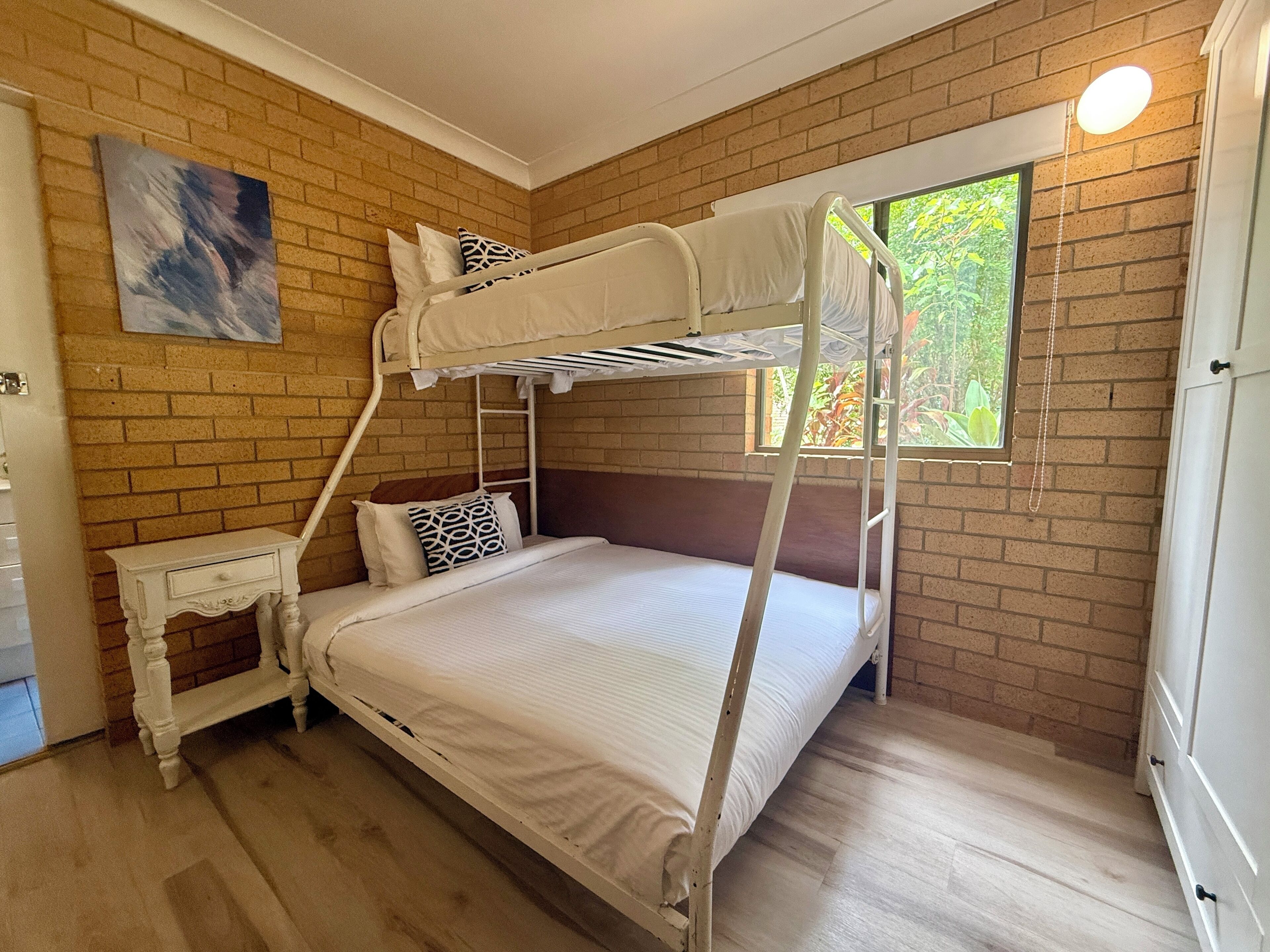 A bunk bed room with a brick wall, a window with a view of greenery, a small bedside table, and a painting on the wall.