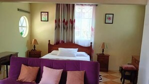 A bedroom with a bed, a purple sofa, two nightstands, a chair, a window with curtains, and a wall with a picture.