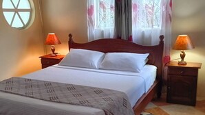 A neatly made bed with white linens and a patterned throw, two bedside lamps, a wooden nightstand, a round window, and a colorful rug.