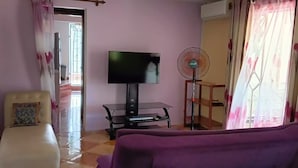 A living room with a purple sofa, a white armchair, a TV on a stand, a standing fan, and a window with curtains.