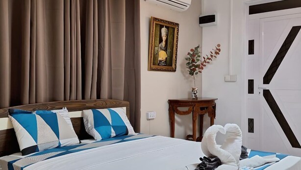 Une chambre d’hôtel avec un lit, des rideaux, un appareil de climatisation, un tableau encadré, une petite table avec un vase et une serviette pliée en forme de cygne.