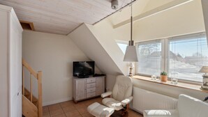 A cozy living room with a TV, a comfortable chair, a sofa, and a window with blinds. - in Dänschendorf auf Fehmarn by Interhome (Bannesdorf)