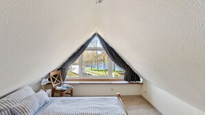 A small, triangular attic bedroom with a bed, pillows, a chair, and a window with curtains. - in Dänschendorf auf Fehmarn by Interhome (Bannesdorf)