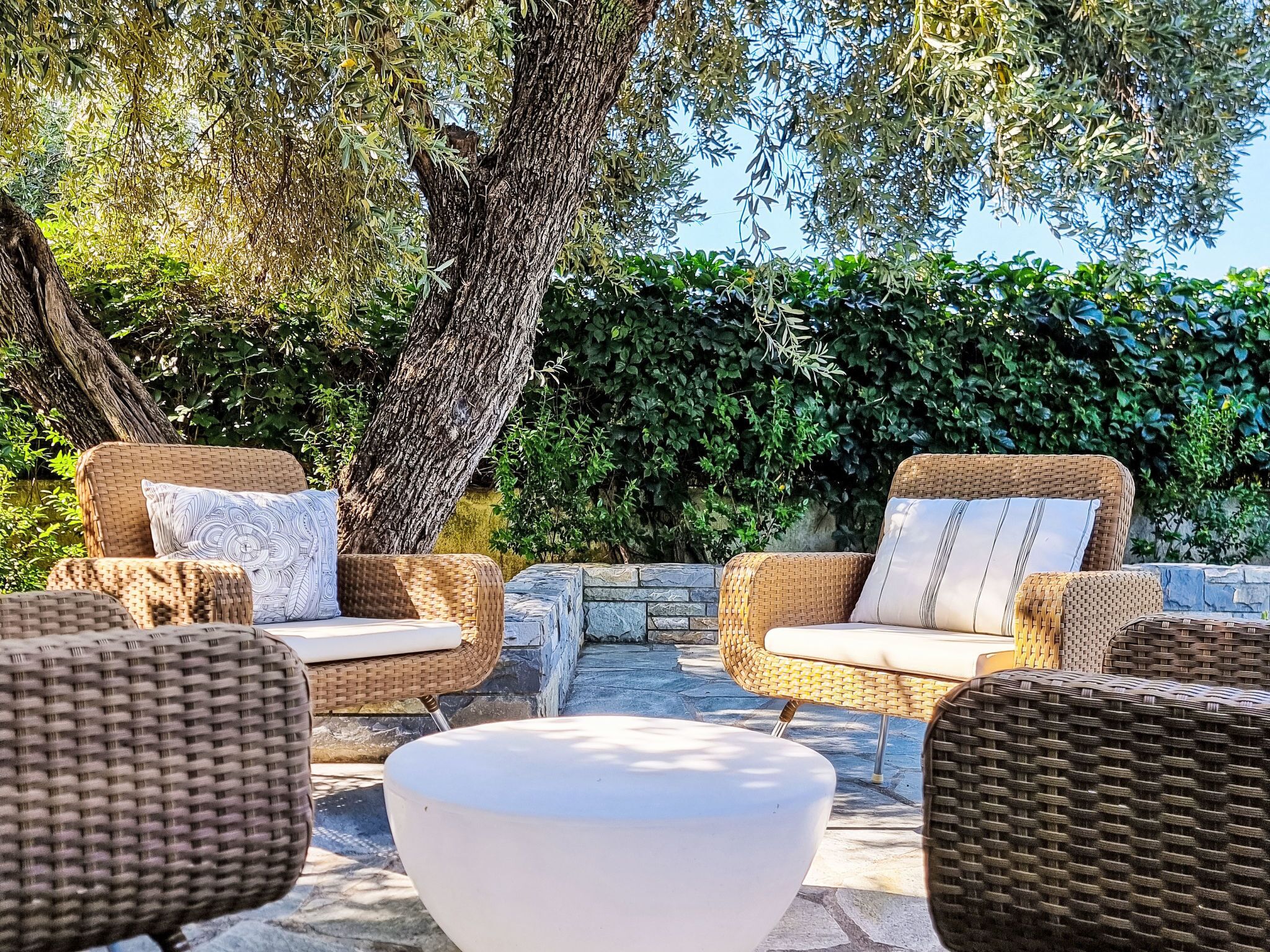 Wicker outdoor furniture with cushions, a stone wall, and a large tree in the background.