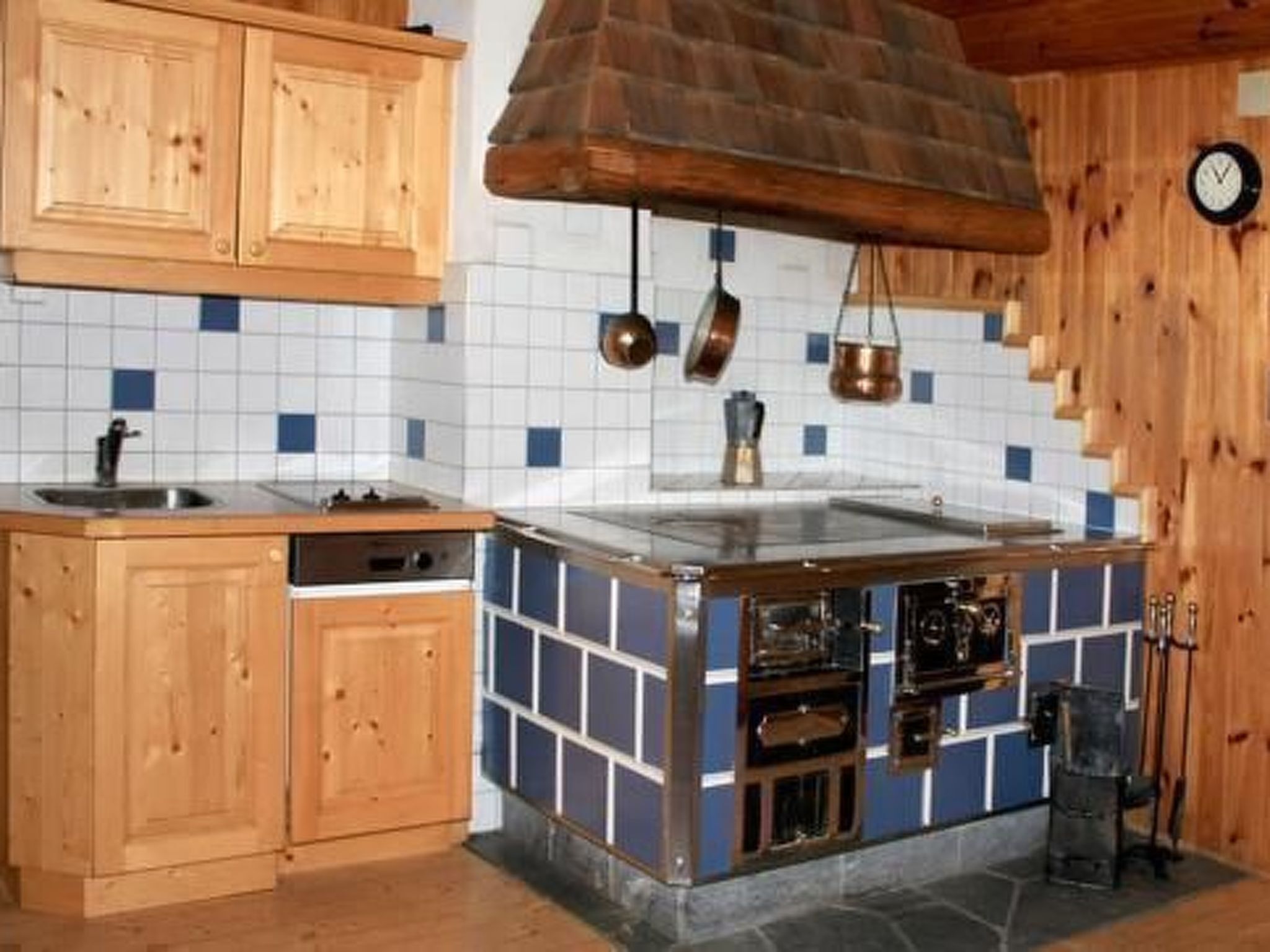 A kitchen with wooden cabinets, a blue and white tiled backsplash, a stove, and hanging copper pots.