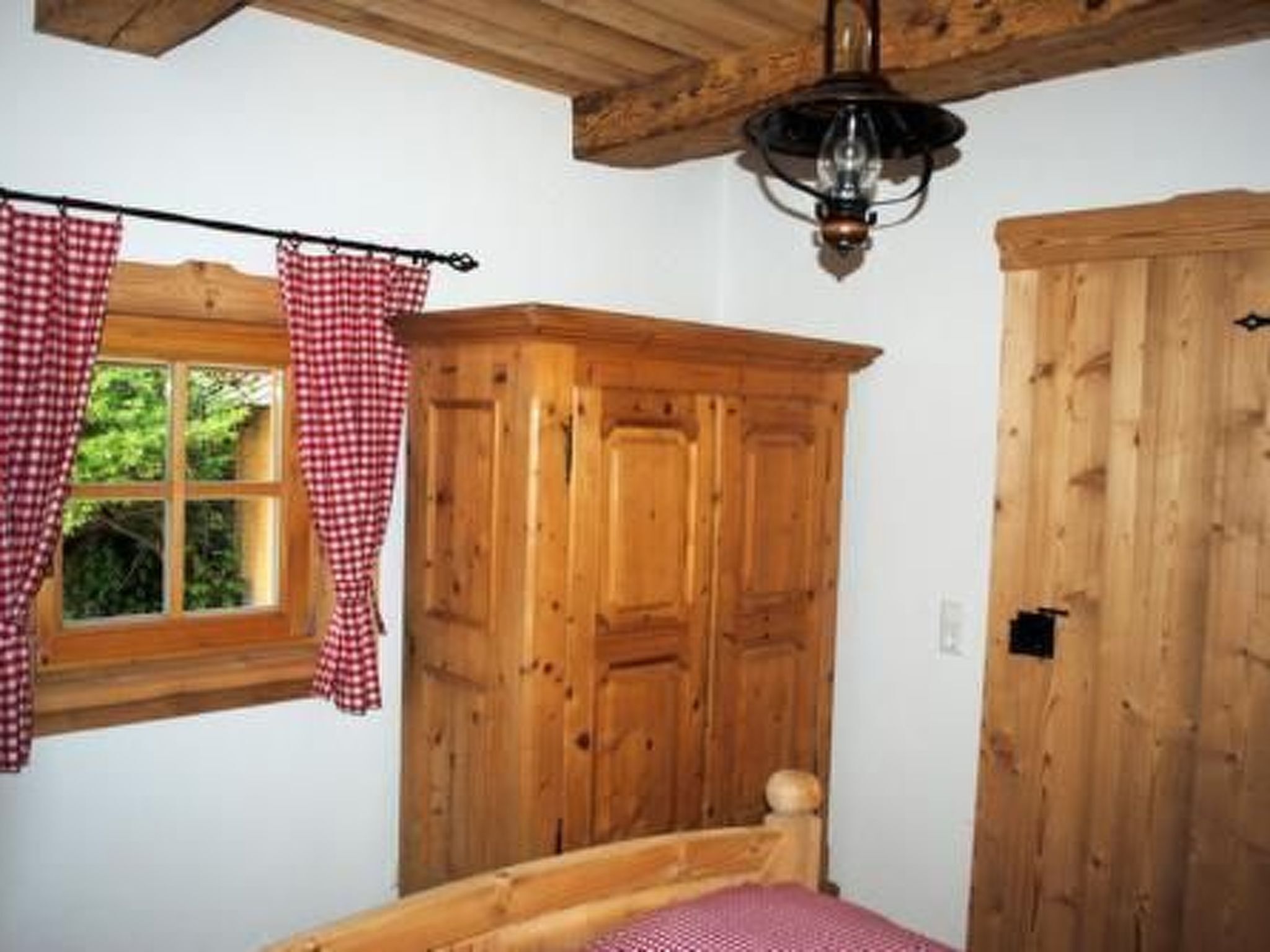 A bedroom with a wooden bed, a window with checkered curtains, and a wooden wardrobe.
