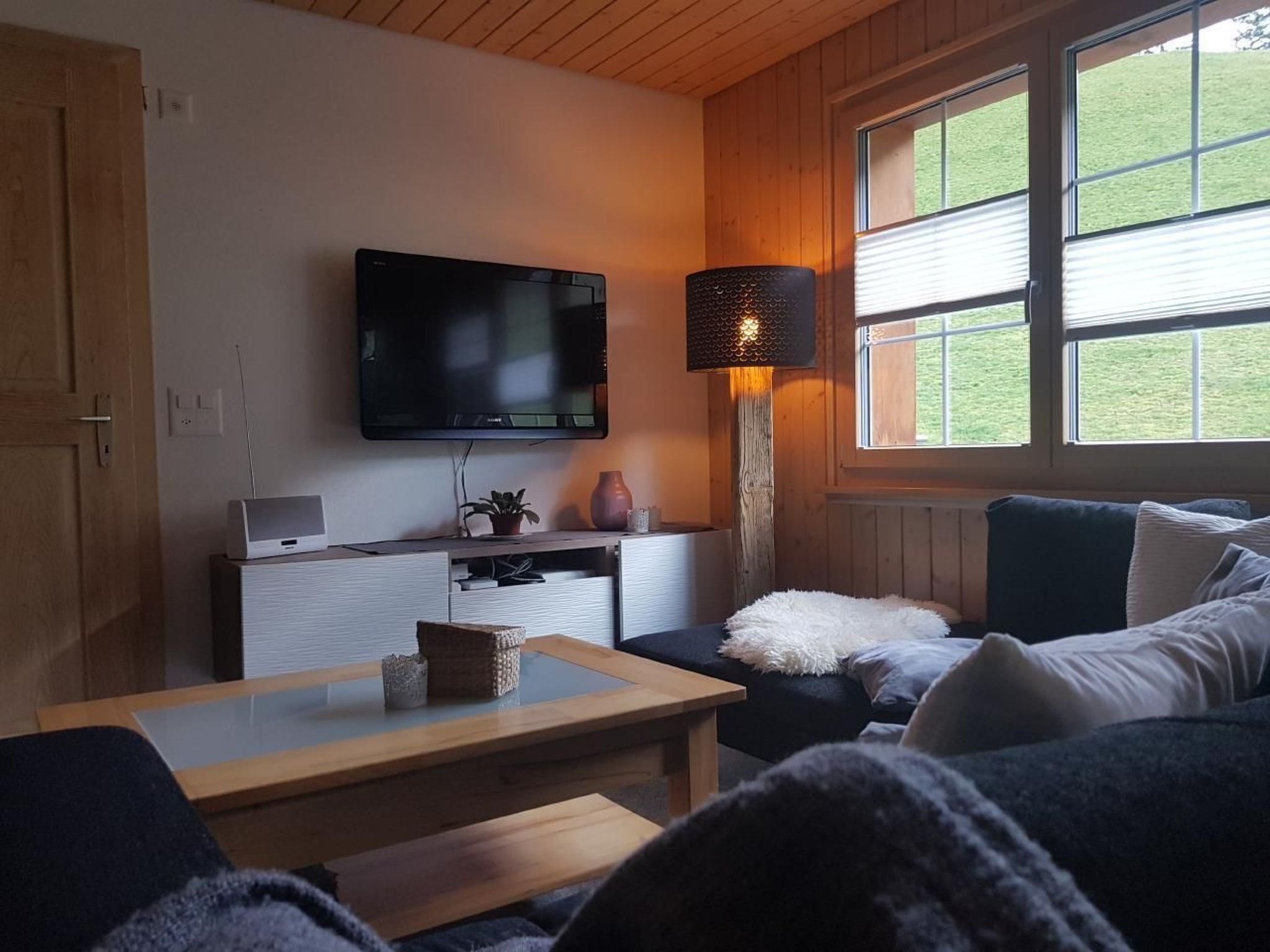 Een gezellige woonkamer met een flatscreen televisie aan de muur, een houten salontafel, een bank met een zachte witte plaid en een vloerlamp.