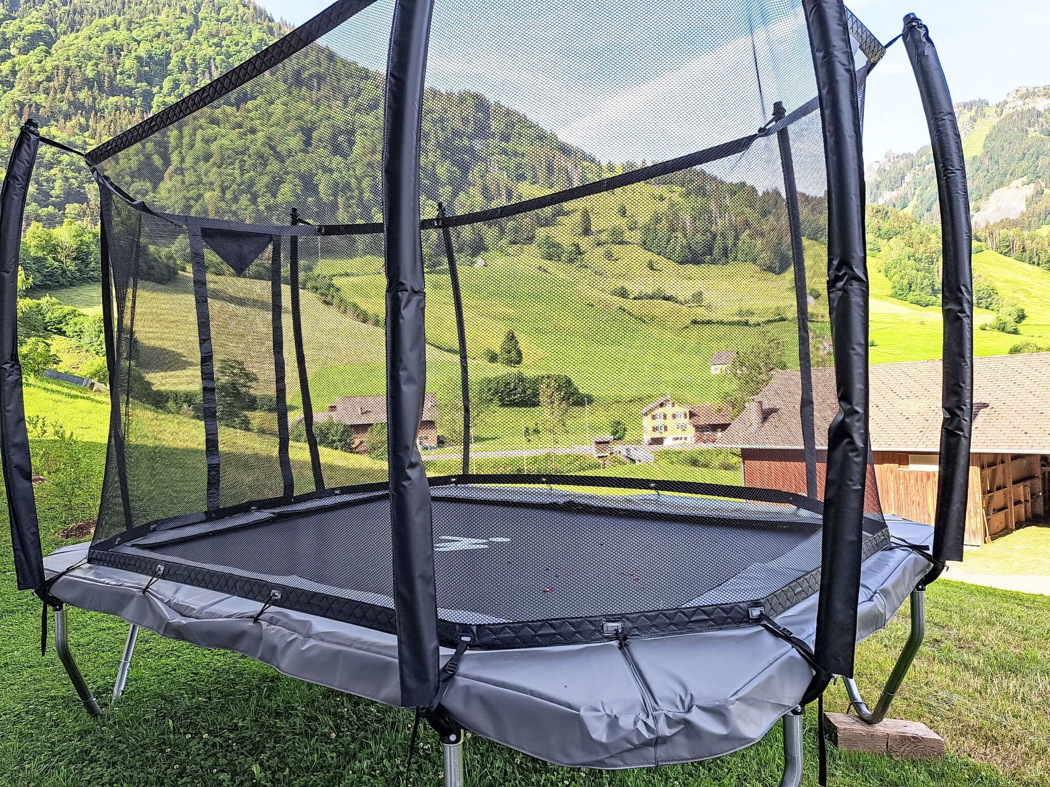 Un trampoline doté d’un filet de sécurité, installé sur une pelouse verdoyante, offrant une vue sur un paysage vallonné et des bâtiments en arrière-plan.
