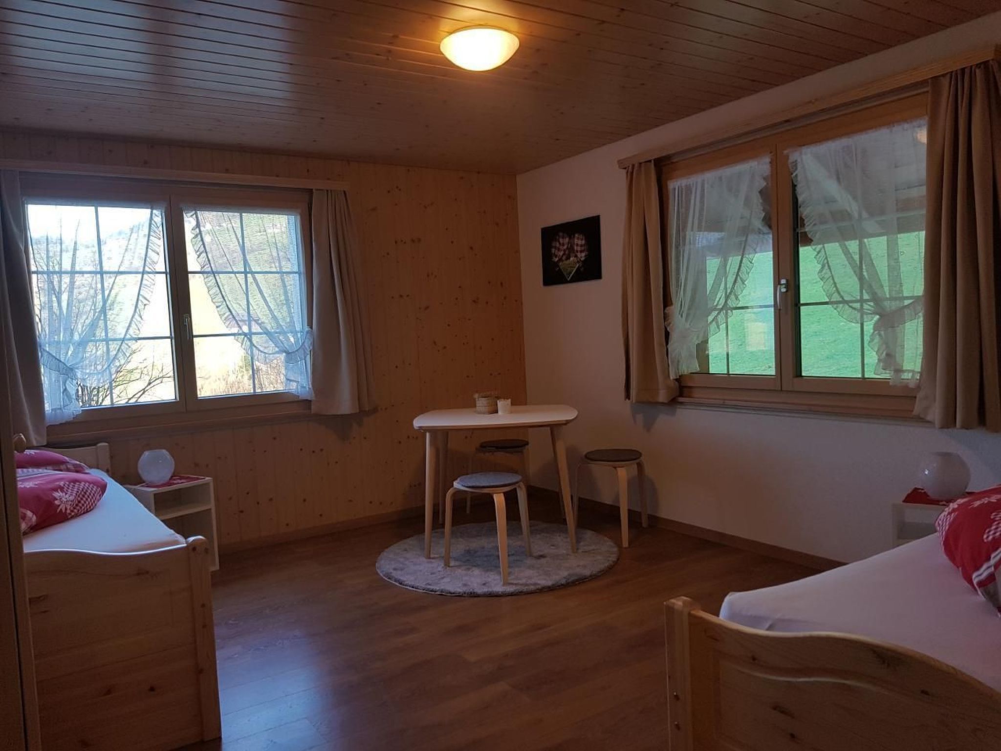 A cozy room with wooden walls and floors, a round table with two stools, a bed with a red pillow, and large windows with curtains.