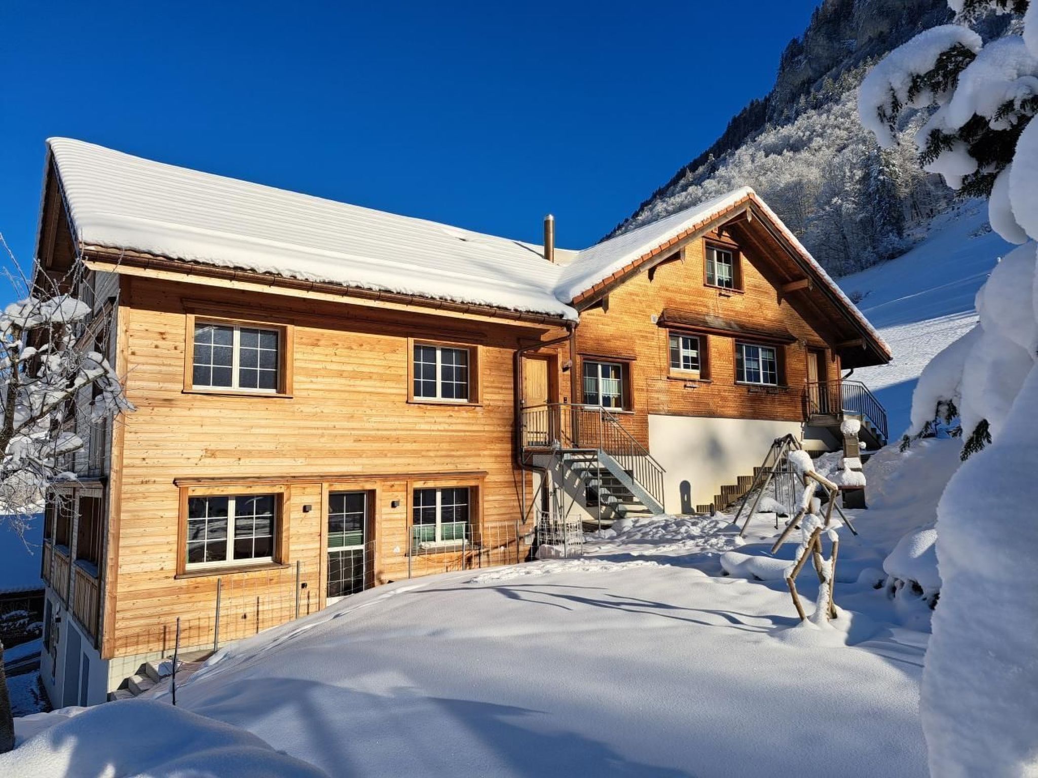 Un chalet en bois doté de nombreuses fenêtres et d’un escalier menant à un balcon, entouré de neige et de montagnes.