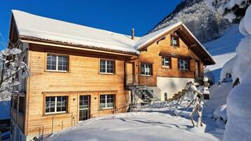 Un chalet en bois doté de nombreuses fenêtres et d’un escalier menant à un balcon, entouré de neige et de montagnes.