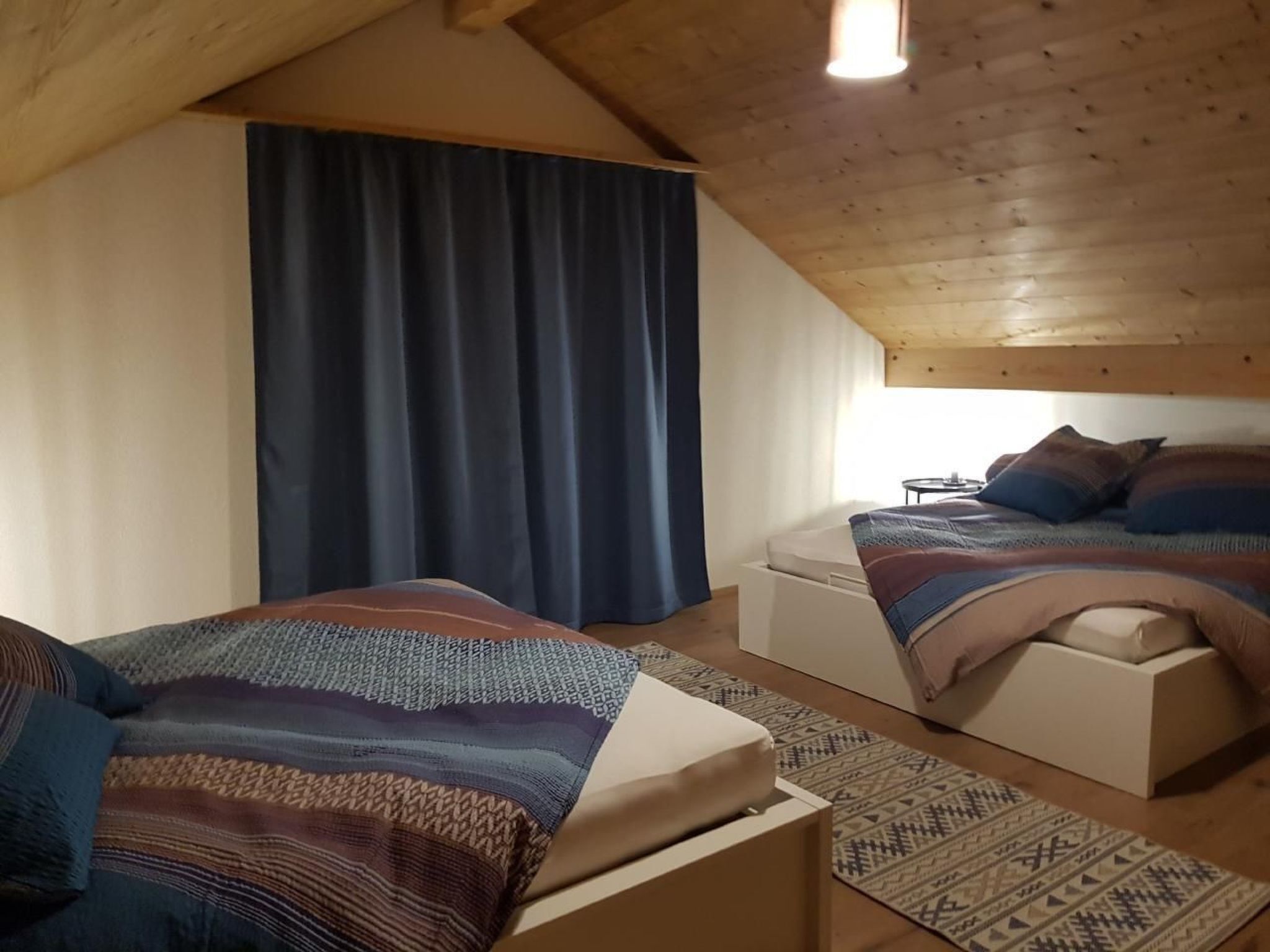 A cozy attic bedroom with two beds, a large window, and a wooden ceiling.