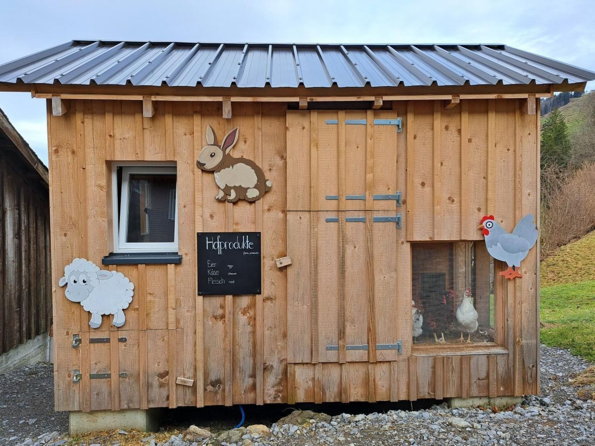 Il s’agit d’une construction agricole en bois, dotée d’un toit en métal, d’une fenêtre et d’une porte. La porte est décorée de motifs représentant des animaux découpés, et une enseigne avec du texte y est fixée.