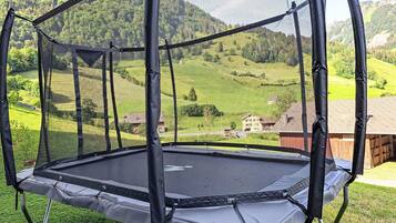 Un trampoline doté d’un filet de sécurité, installé sur une pelouse verdoyante, avec un paysage montagneux en arrière-plan.