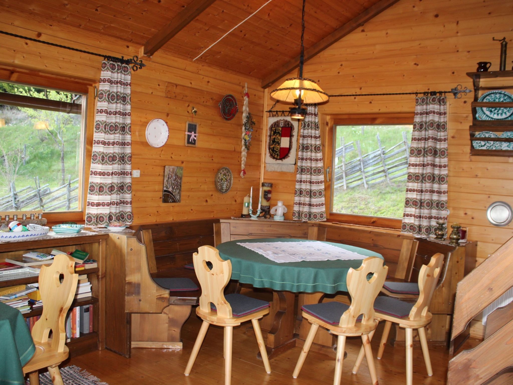 A wooden cabin with a dining area featuring a table and chairs, a bookshelf, and a window with curtains.