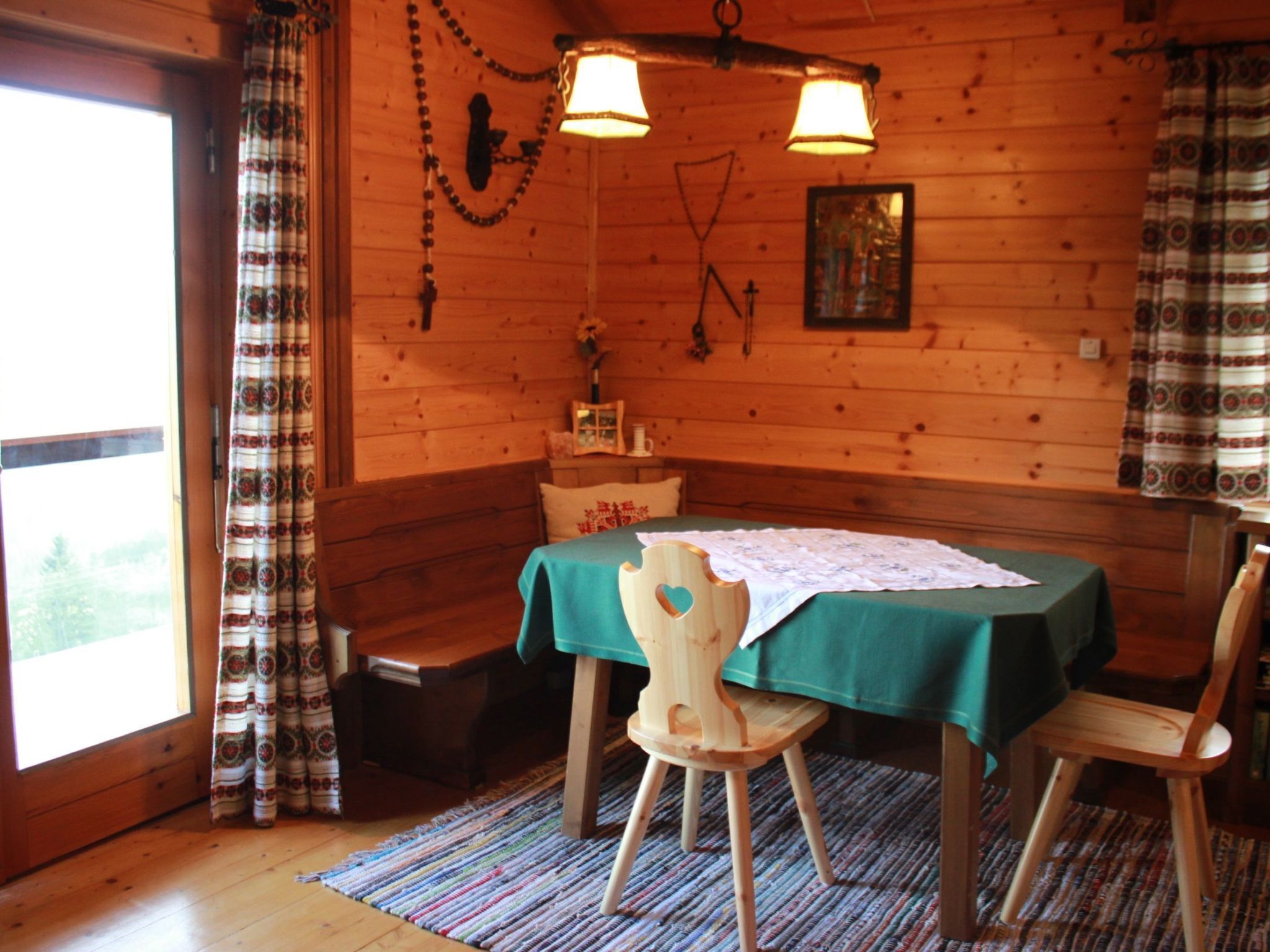 Ein Interieur einer Holzhütte mit Esstisch, zwei Stühlen, einer Bank und einem Fenster mit Vorhängen.