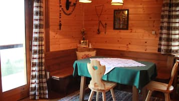 Ein Interieur einer Holzhütte mit Esstisch, zwei Stühlen, einer Bank und einem Fenster mit Vorhängen.