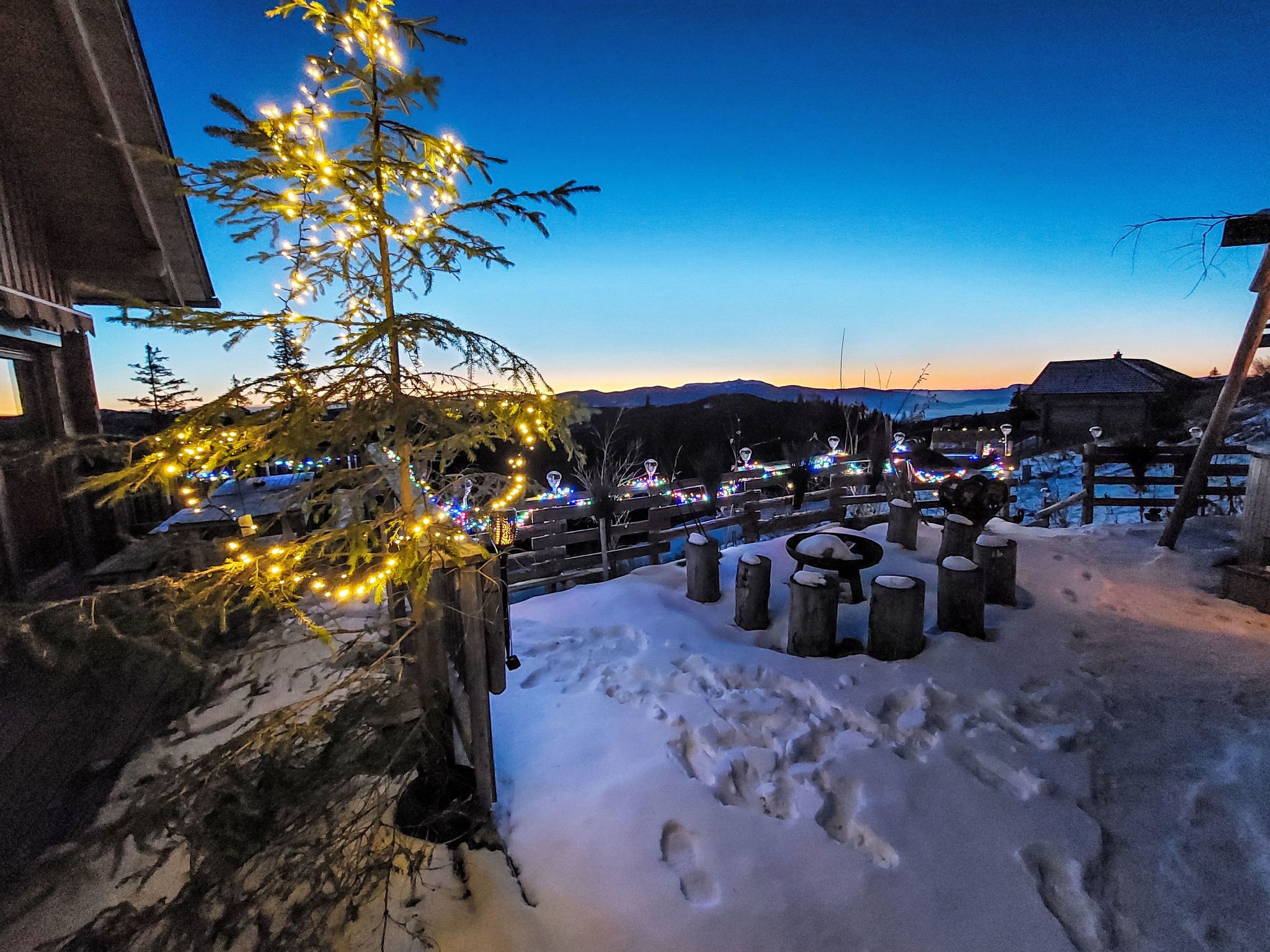 Un espacio exterior cubierto de nieve, con una valla de madera, suelo nevado y un árbol con luces.