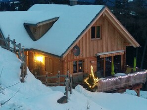 A wooden cabin with a sloped roof, a porch with lights, and a snow-covered landscape.