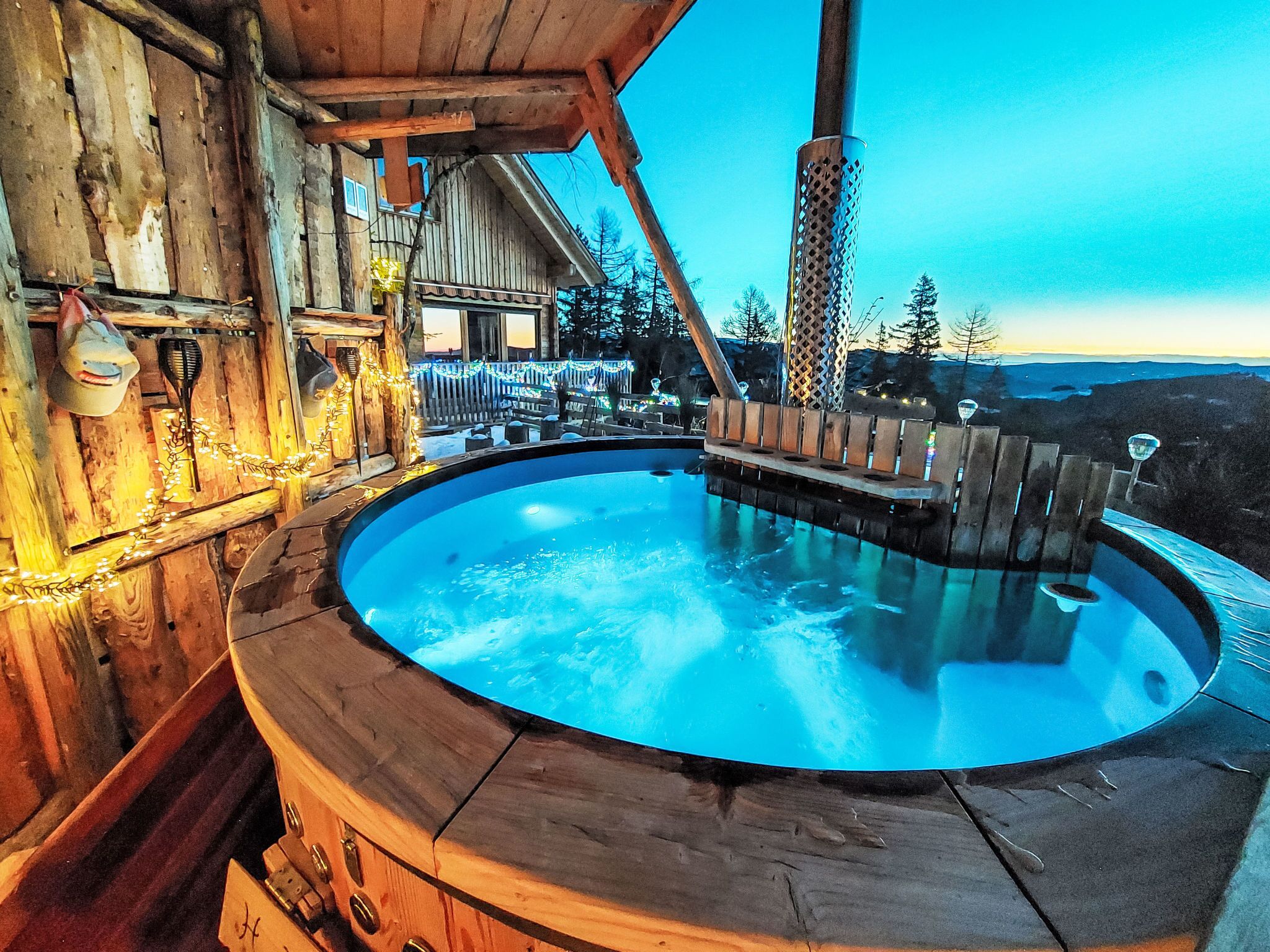A wooden hot tub with a wooden deck surrounding it, set against a backdrop of a wooden cabin and a clear sky.