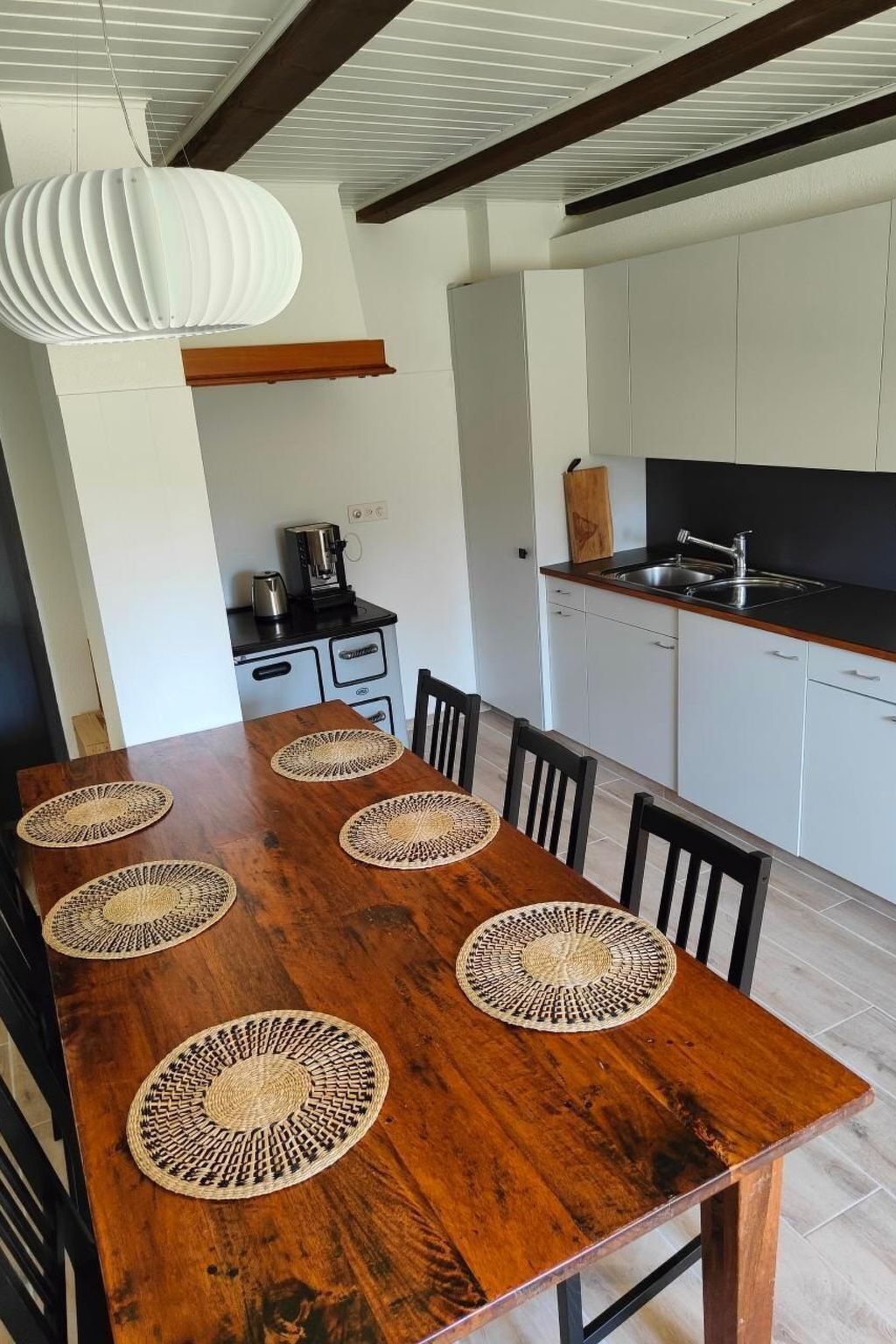 A dining area with a wooden table, six woven placemats, black chairs, a hanging light fixture, a kitchen with white cabinets, and a black countertop.