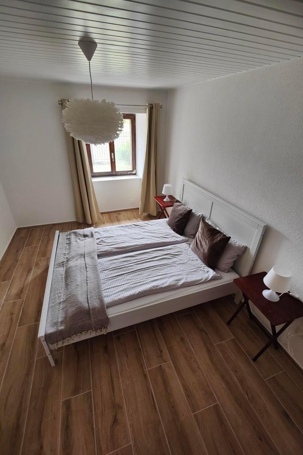 A bedroom with a large bed, a bedside table with a lamp, a window with curtains, and a hanging light fixture.