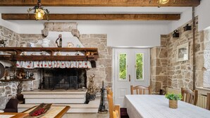 Ein rustikaler Innenraum mit Kamin aus Stein, Holzbalken und einer Essecke mit Tisch und Stühlen.