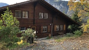 Un edificio de madera con múltiples ventanas y techo inclinado, rodeado de árboles y montañas al fondo.
