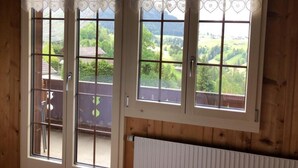 Una habitación con tres ventanas, cortinas de encaje con forma de corazón, vistas a un paisaje verde y decoración de madera.