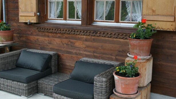 Pared exterior de madera con una ventana, un sofá de mimbre con cojines negros y plantas en macetas.