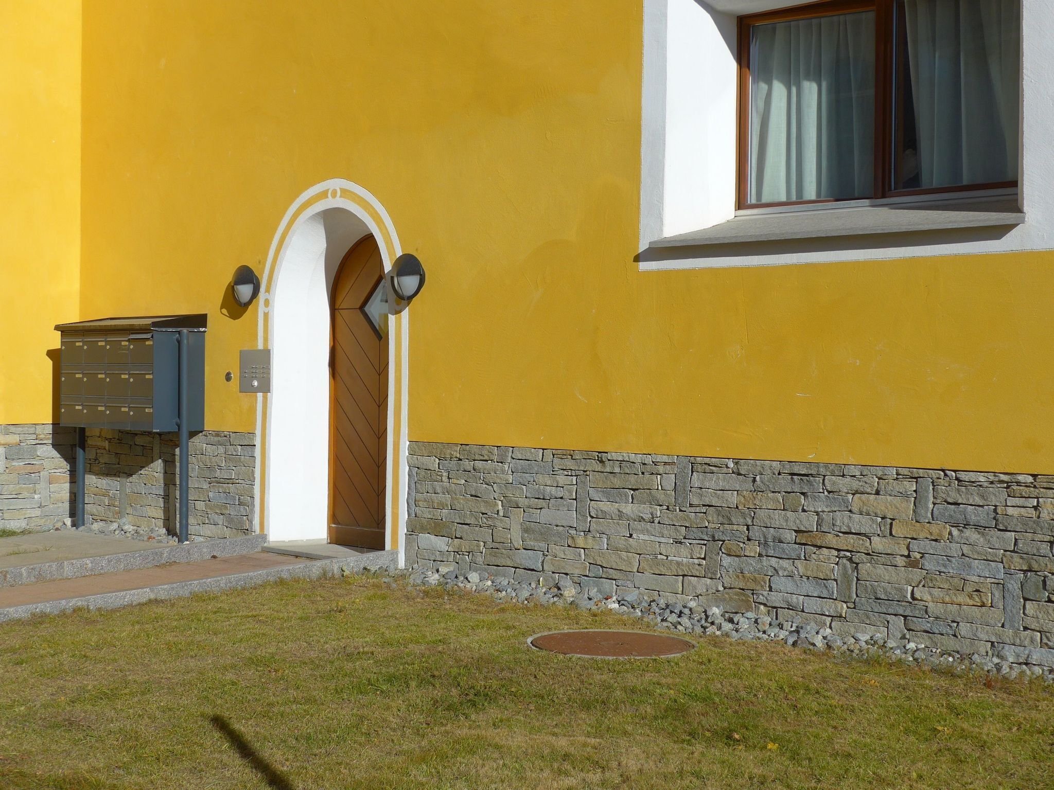 Ein gelbes Gebäude mit steinernem Fundament, einem Briefkasten und einer Tür mit Bogenöffnung.