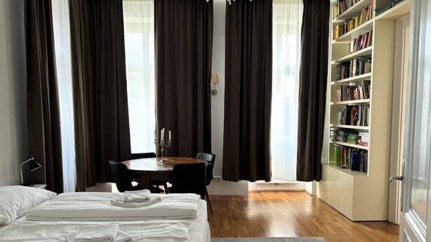 Habitación de hotel con una cama grande, una mesa de comedor con sillas, una estantería y un candelabro.
