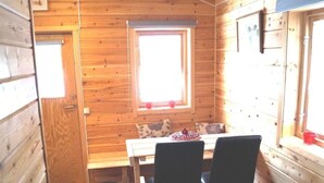 Una pequeña cabaña de madera con un ventanal, una mesa, dos sillas y un bote de basura negro.
