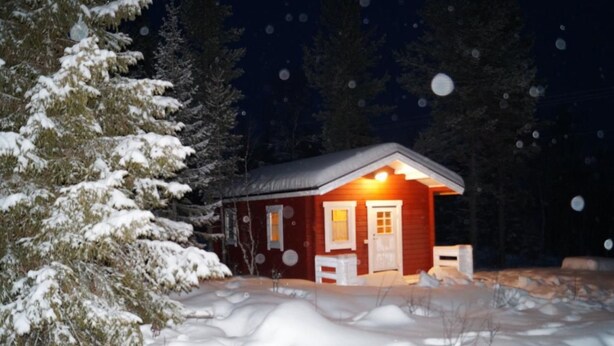 Una cabaña con fachada roja, puerta blanca y una ventana iluminada, rodeada de nieve y árboles.
