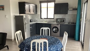 Ett kompakt kök med mörka skåp, en tvättmaskin, en mikrovågsugn och en matplats med ett bord och stolar.