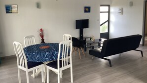 En matplats med ett runt bord, vita stolar, en svart soffa, en tv och ett fönster som släpper in naturligt ljus.