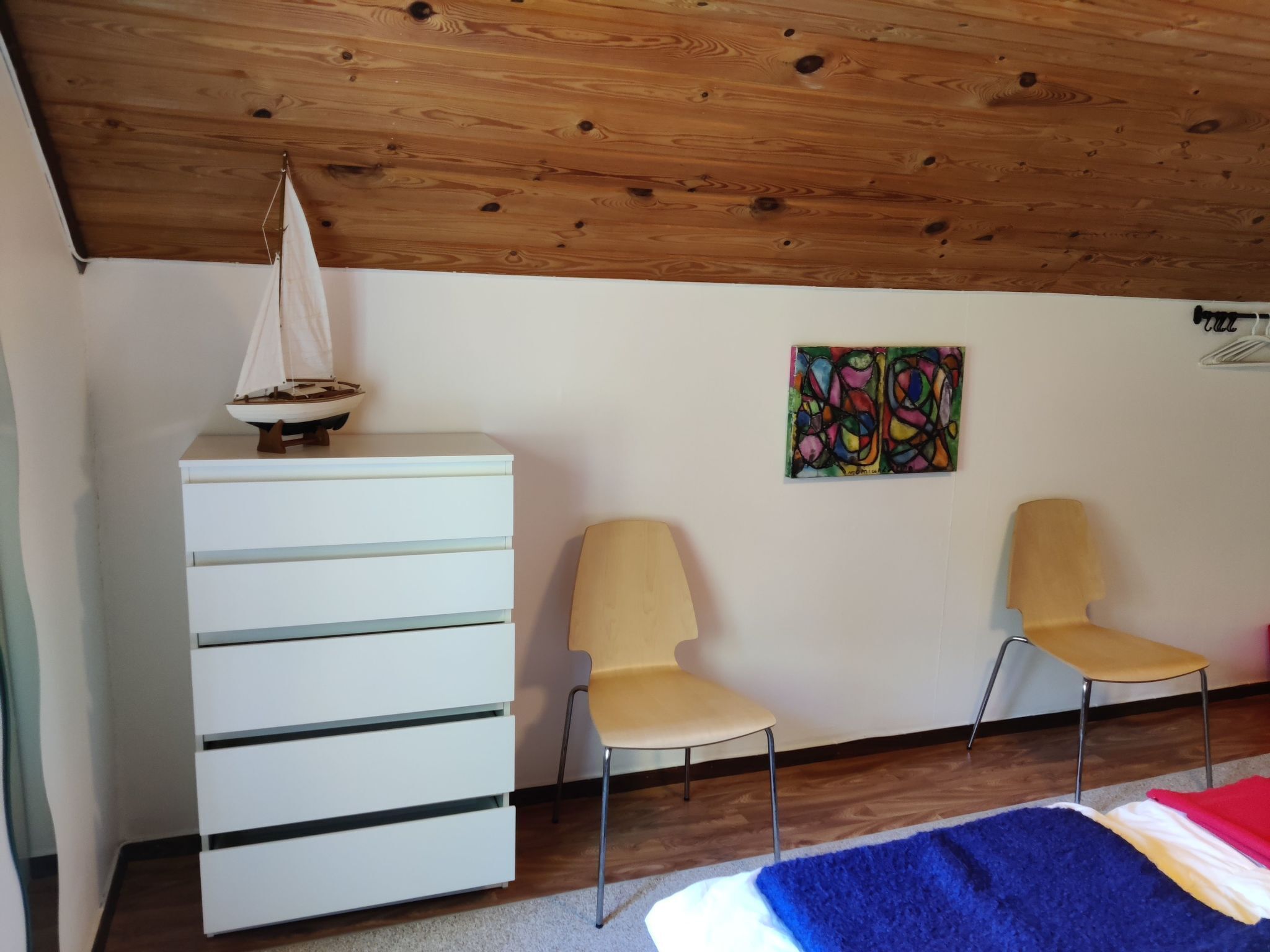 Una stanza con soffitto in legno, una cassettiera bianca, due sedie, un quadro astratto dai colori vivaci e un modellino di barca a vela.