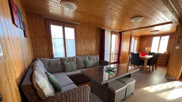 L’intérieur chaleureux d’un chalet avec un grand canapé à ossature en bois, une table à manger avec un plateau en verre et des chaises en osier.