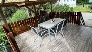 Un patio couvert avec une table et des chaises, entouré d’une clôture en bois et offrant une vue sur un bâtiment en arrière-plan.