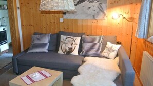 A cozy room with a wooden wall, a grey sofa with patterned cushions, a white furry throw, a small table with a placemat, and a mountain painting above the sofa.