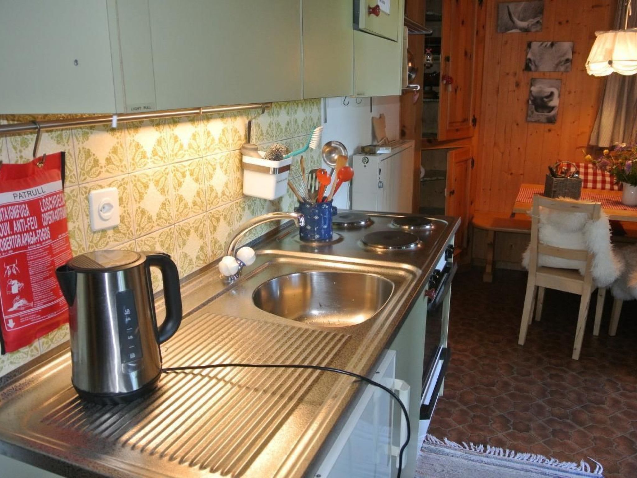Una cocina con un lavamanos de acero inoxidable, una tetera y una estufa. Hay un armario de madera sobre el lavamanos y un refrigerador pequeño a la derecha.