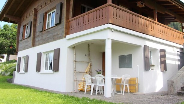 A two-story building with a balcony, white exterior walls, and a covered outdoor seating area with chairs and a table.