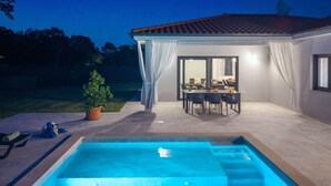 Ein moderner Poolbereich im Freien mit einer überdachten Terrasse, einem Esstisch und Blick in ein Wohnzimmer.