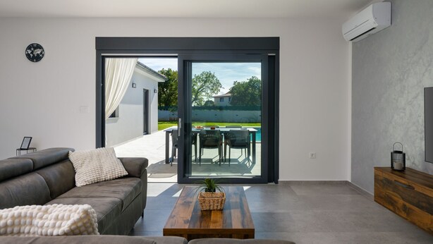 Ein modernes Wohnzimmer mit einer grauen Couch, einem Holztisch und einer großen Schiebeglasdoor zum Außenbereich mit Pool und Essbereich.