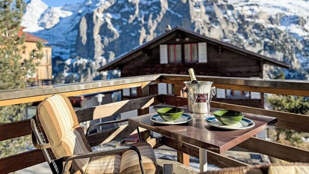 A table with a bottle of champagne, two green bowls, and a wooden chair with striped cushions. A mountain range with snow-covered peaks is visible in the background. - Rottal 4 Bett Wohnung by Interhome (Lauterbrunnen)