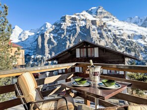 A table with a bottle of champagne, two green bowls, and a wooden chair with striped cushions. A mountain range with snow-covered peaks is visible in the background. - Rottal 4 Bett Wohnung by Interhome (Lauterbrunnen)