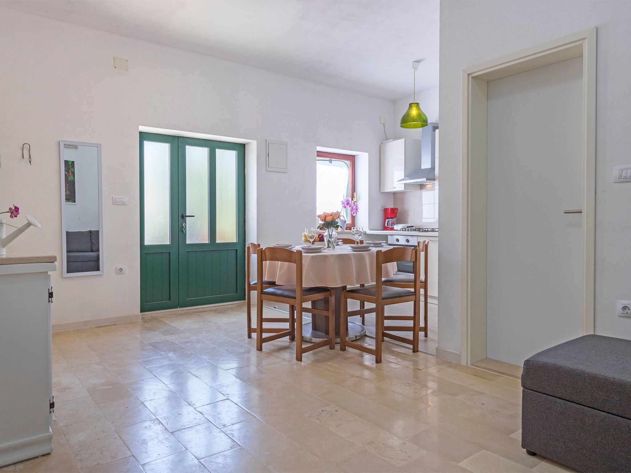 A dining area with a table set for four, a green door, and a kitchen with a stove and a green pendant light.