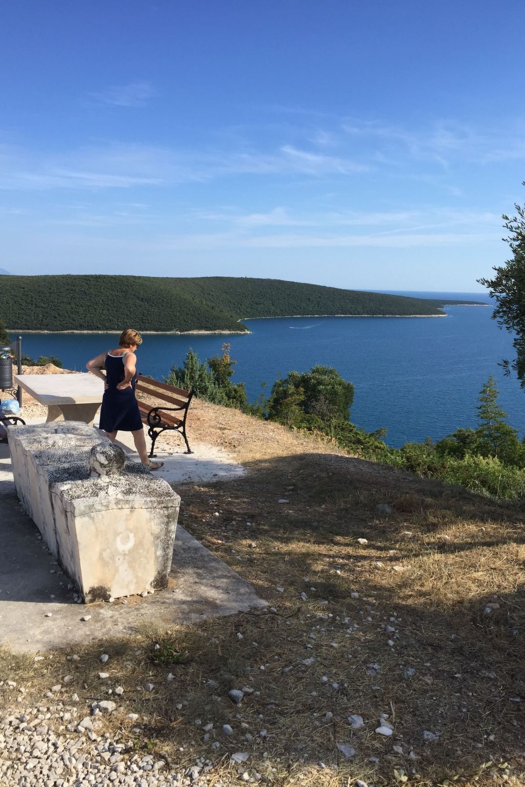 A person standing by a concrete bench overlooking a body of water with a forested hill in the background.