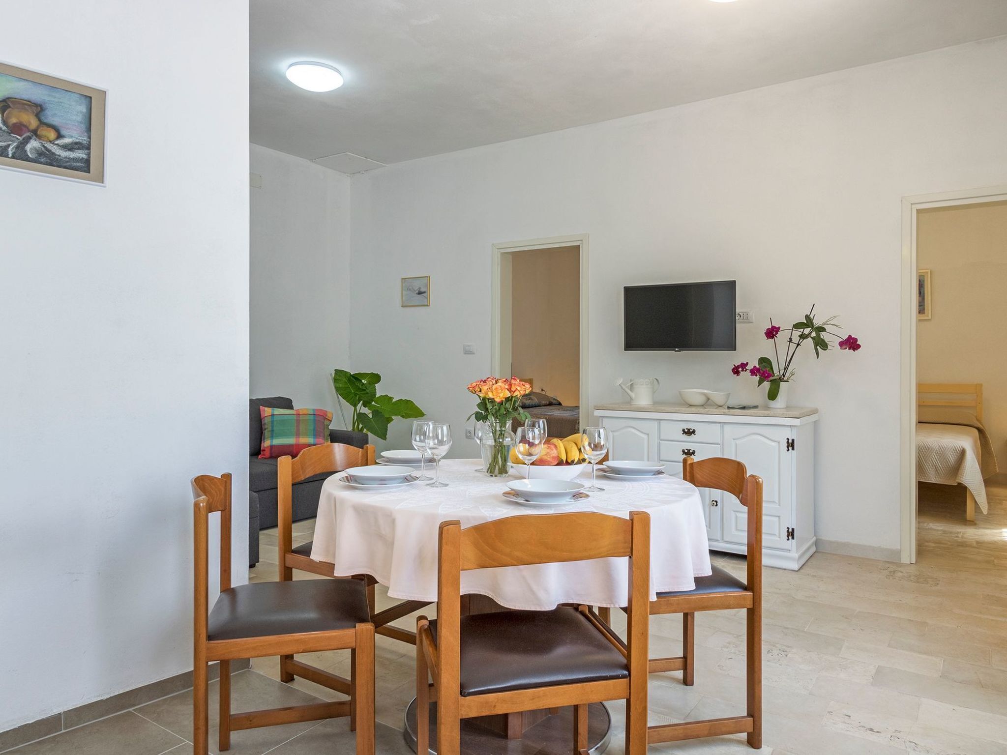 A dining area with a round table set for four, surrounded by wooden chairs. A television is mounted on the wall above the table.